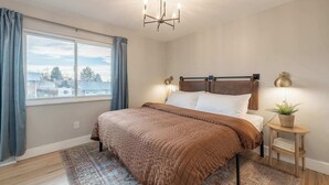A bedroom with a large bed, a window with curtains, a chandelier, a bedside table with a lamp, and a small potted plant. - Denver Retreat • Hot tub+Firepit+Games • Fast Wifi (DENVER)