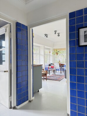 Un couloir aux murs recouverts de carreaux bleus, une porte blanche et une vue sur une pièce où l’on aperçoit un tapis à motifs, une chaise et une table.