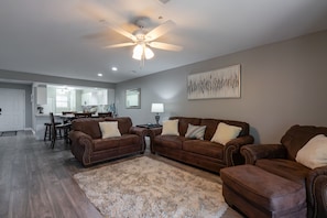 A spacious living room with two brown leather sofas, a large fluffy rug, a ceiling fan, and a dining area in the background.