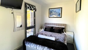 Une chambre à coucher comprenant un lit, une table de chevet, une commode, un téléviseur fixé au mur et une fenêtre avec des rideaux.