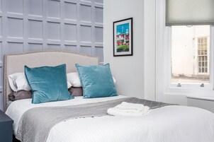 A neatly made bed with a striped bedspread and two teal pillows, a framed picture on the wall, and a window with a view of buildings.
