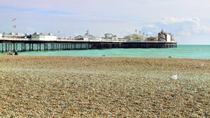 A pebbled beach with a pier extending into the sea, featuring buildings and a lighthouse. - Studio In the Lanes (Brighton)