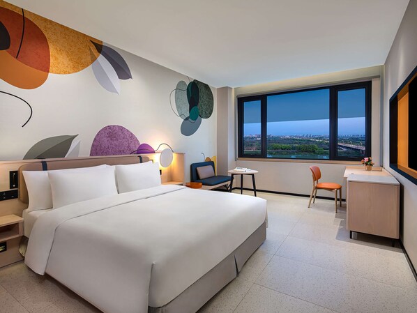 A modern hotel room with a large bed, a desk, a chair, and a view of the city through a large window.