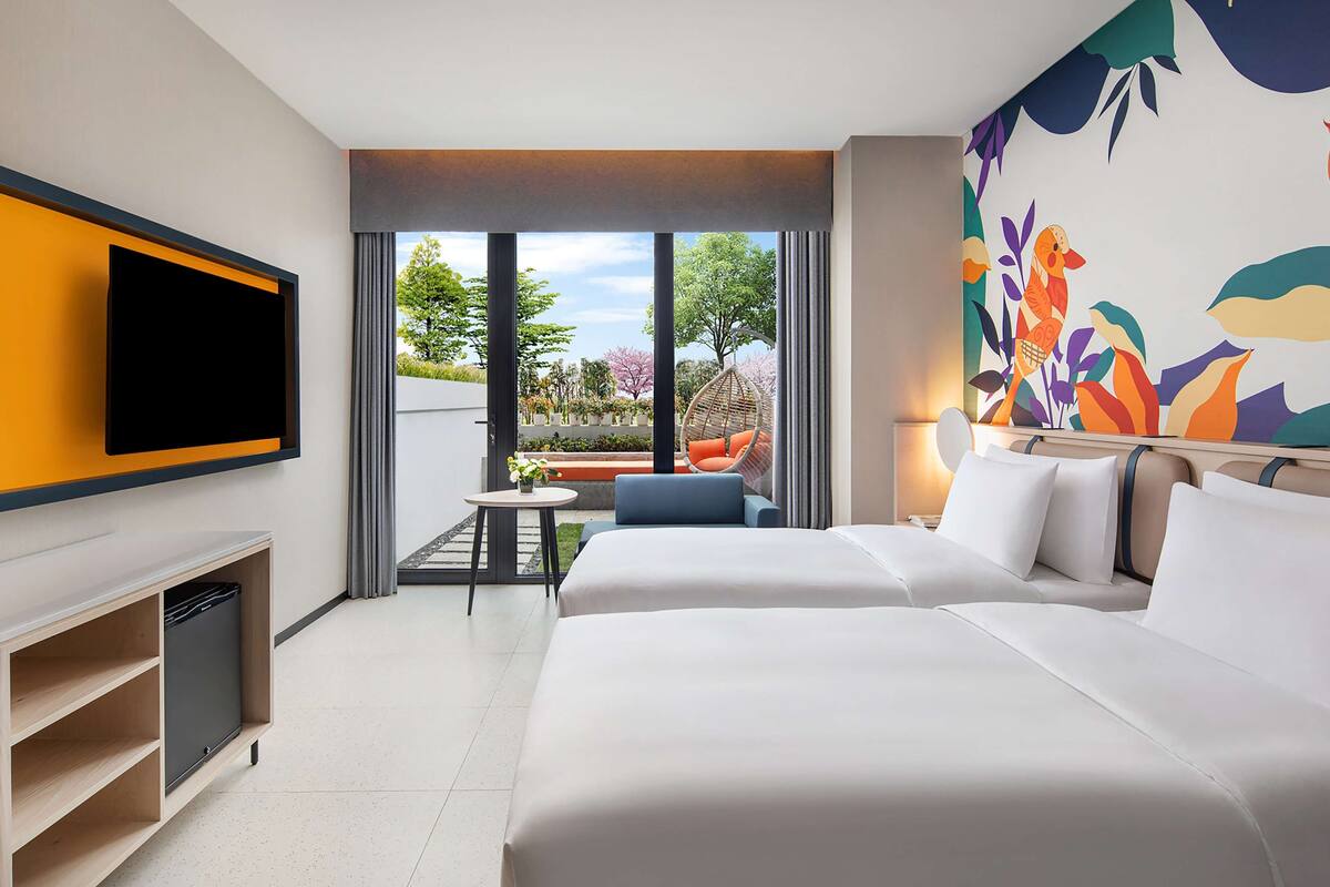 Une chambre d’hôtel moderne dotée d’un grand lit, d’un téléviseur à écran plat fixé au mur, d’une petite table avec une chaise et d’un balcon offrant une vue sur la verdure.