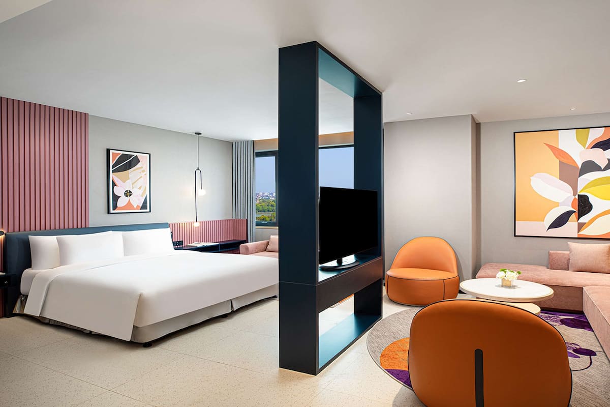 Une chambre d’hôtel moderne avec un grand lit, une télévision, un canapé et un fauteuil. Il y a une grande fenêtre et un tableau au mur.