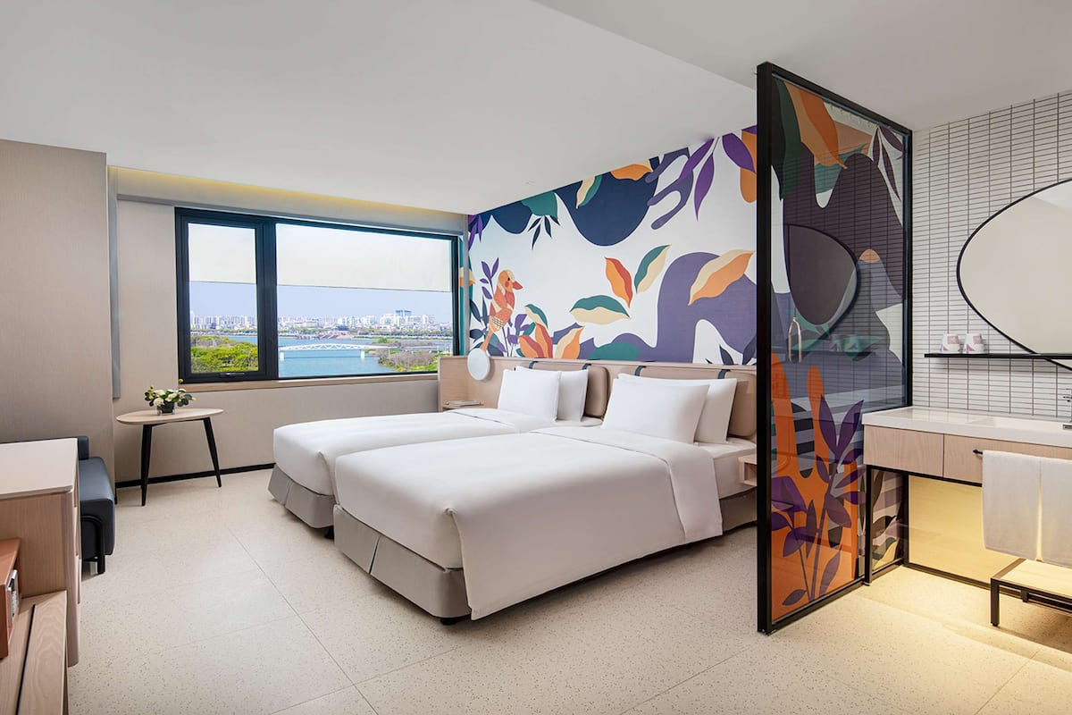 Une chambre d’hôtel moderne, dotée d’un grand lit, d’un bureau, d’une chaise, d’une petite table, d’un miroir et offrant une vue sur la ville par la fenêtre.