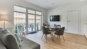 A modern living room with a grey sofa, a dining table with chairs, a TV mounted on the wall, and a sliding glass door leading to an outdoor balcony. - Apartamenty Swinoujscie – Lissa (Swinoujscie)