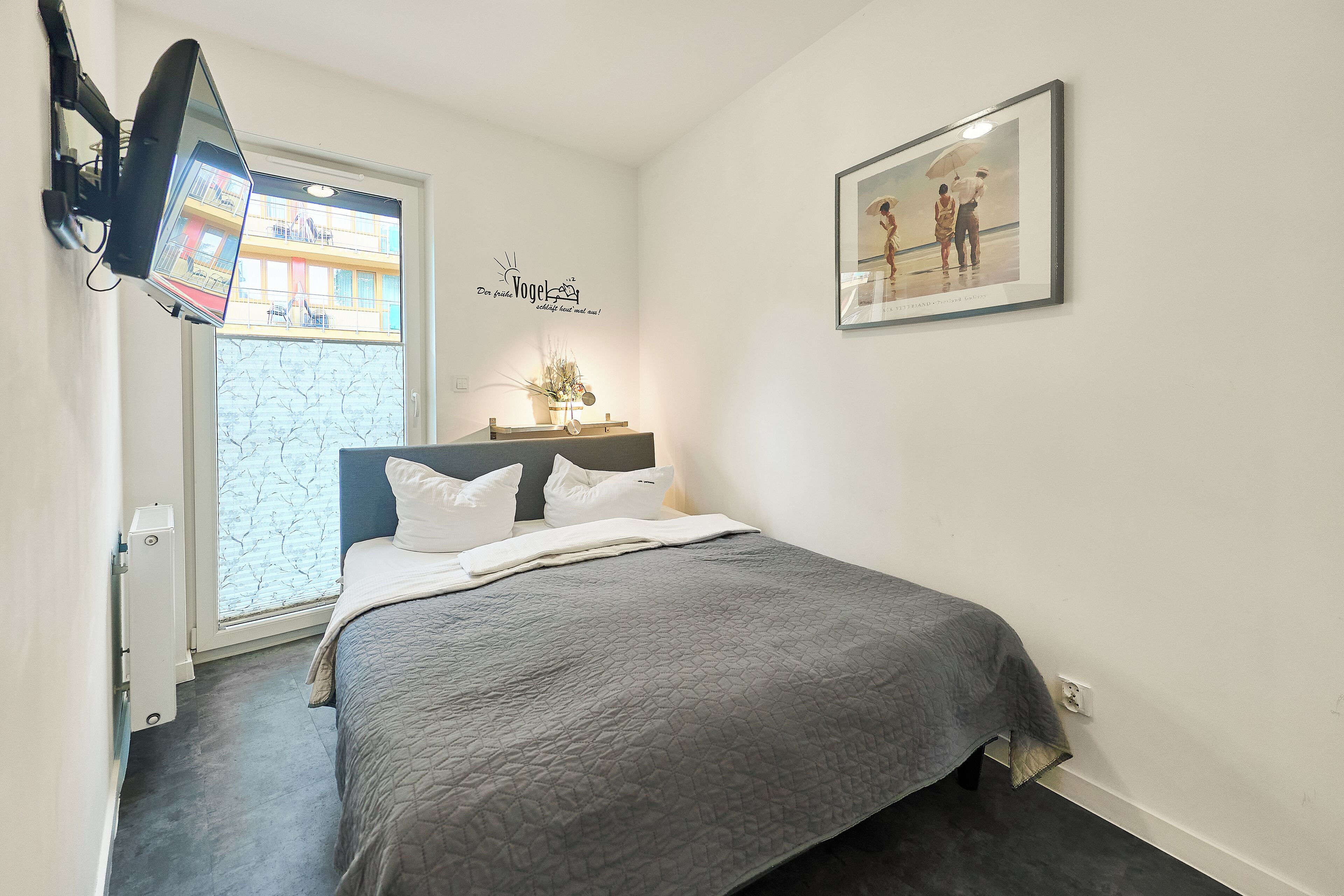 A bedroom with a bed, a window, a wall-mounted TV, and a framed picture.
