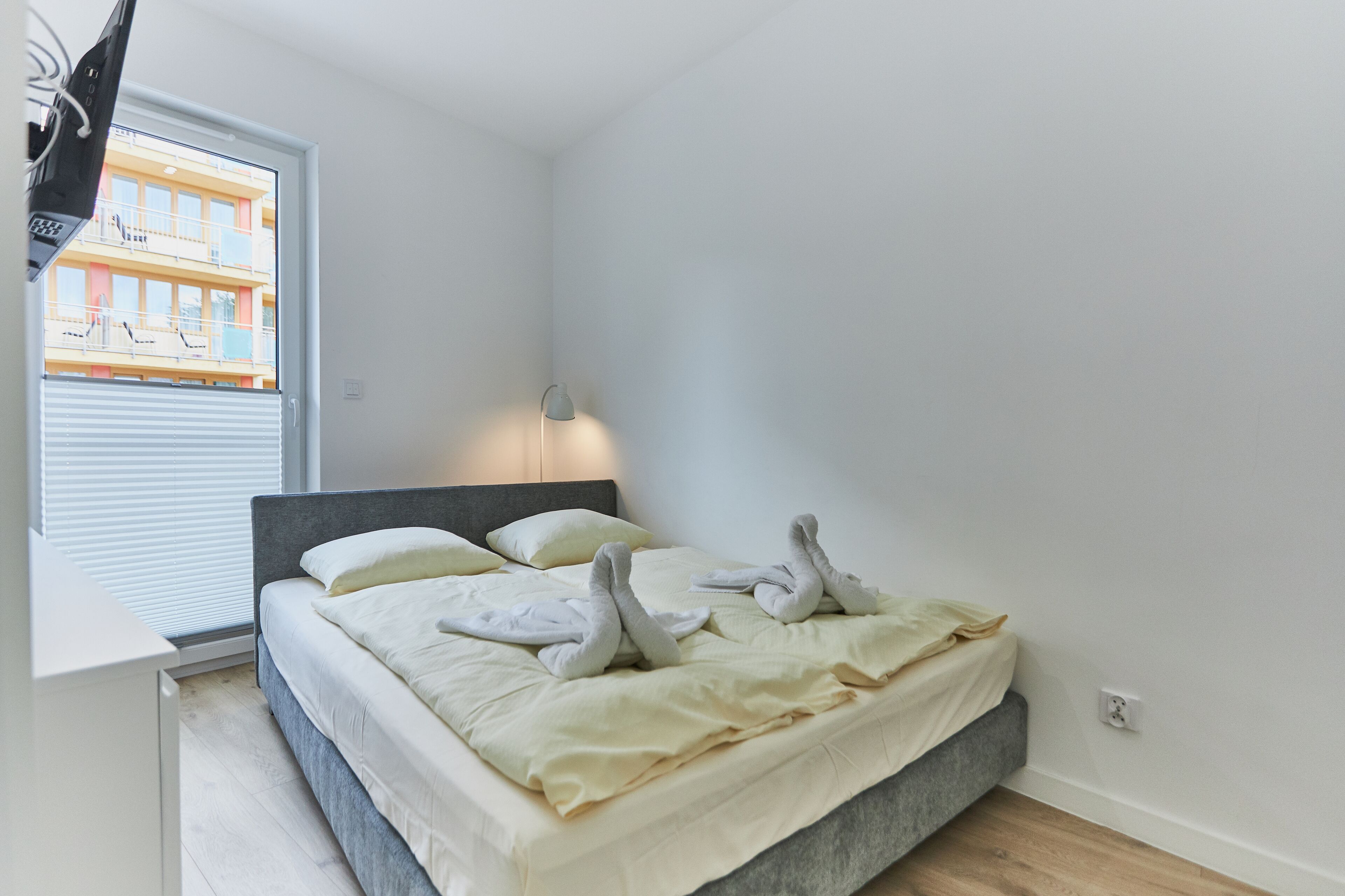 A modern bedroom with a bed, a bedside lamp, and a view of a building through a window.