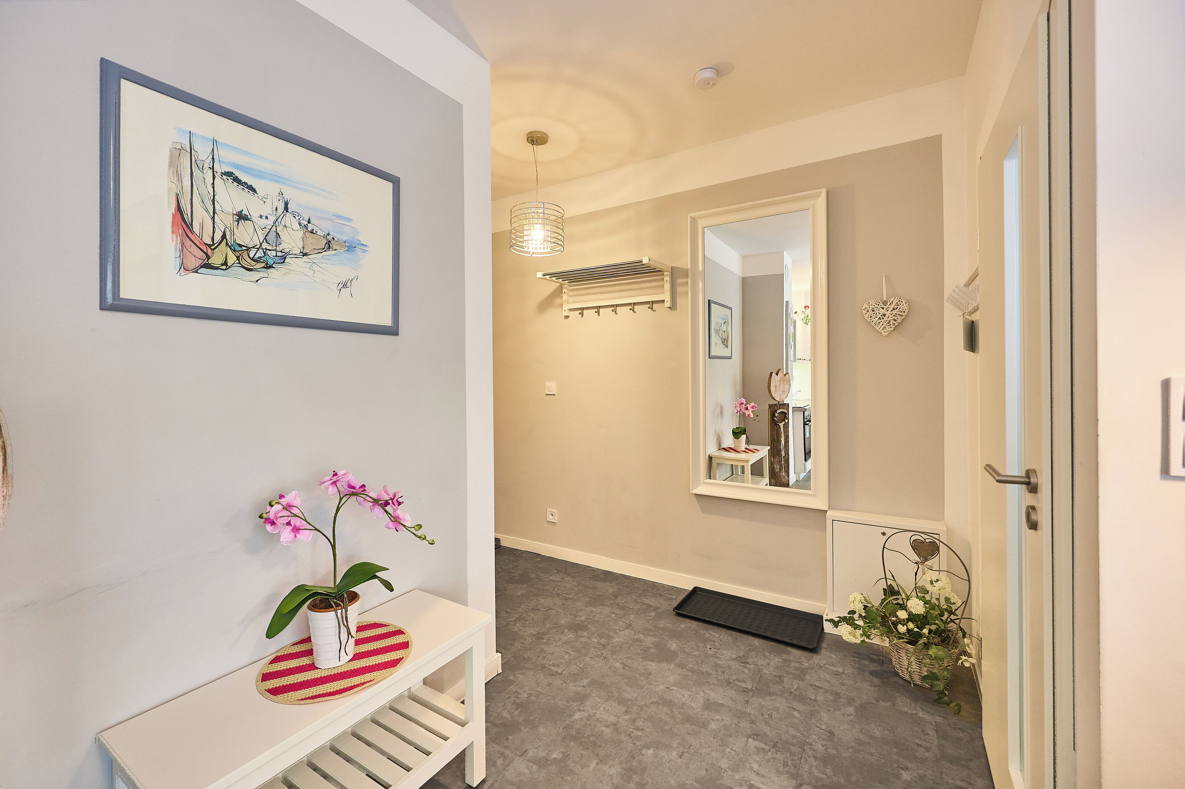 A hallway with a framed artwork, a potted orchid, a white bench, a mirror, and a door.