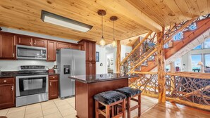 A kitchen with wooden cabinets, stainless steel appliances, and a central island with bar stools. A wooden staircase with a decorative railing leads to an upper level. - Upscale Home With Expansive Mountain Views (Banner Elk)