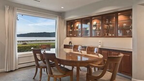 Un coin repas comprenant une table ronde et six chaises, une grande fenêtre avec des rideaux et un meuble contenant de la verrerie.