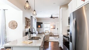 A modern kitchen with a central island, stainless steel appliances, pendant lighting, and a view into the living area.
