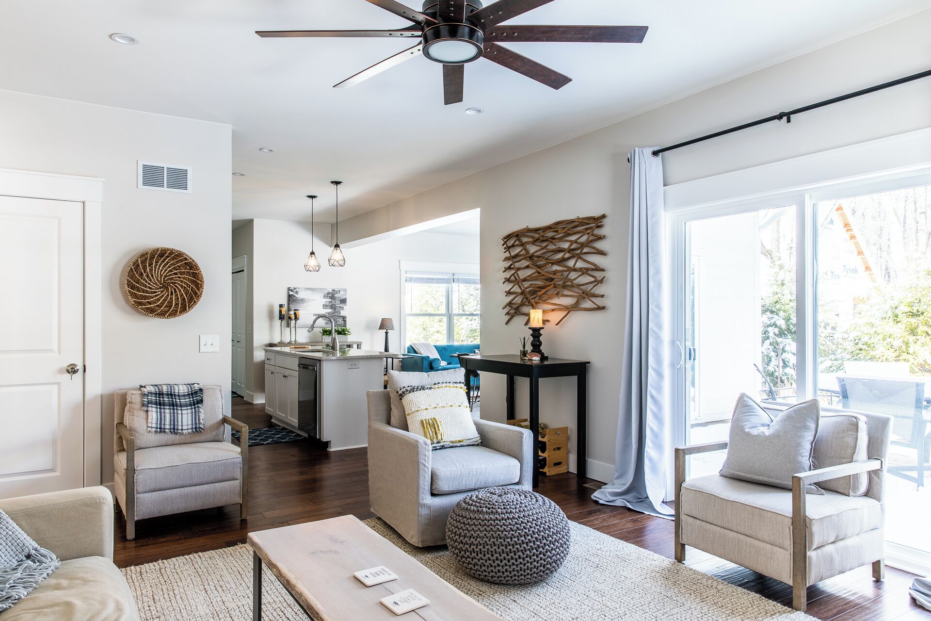 A spacious living room with a large window, a ceiling fan, and a kitchen area in the background.