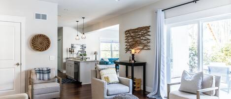 A spacious living room with a large window, a ceiling fan, and a kitchen area in the background.