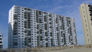 Un immeuble résidentiel de grande hauteur, doté de nombreux balcons, avec un rez-de-chaussée sablonneux et un ciel dégagé.