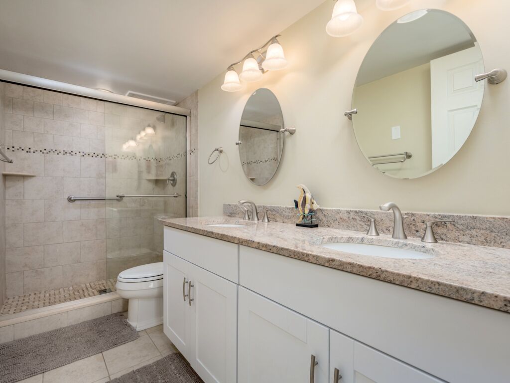 Une salle de bain moderne dotée d’une douche à l’italienne, de toilettes, d’un meuble-lavabo à deux vasques et de deux miroirs fixés au mur.