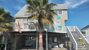 A beachfront property with a covered patio, outdoor seating, and a staircase leading to the entrance. - Cozy 3BR oceanfront home in Holden Beach, perfect for family getaways. (Holden Beach)