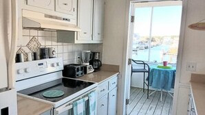 A compact kitchen with white cabinets, a stove, and a toaster. A balcony with a table and chairs is visible through a glass door. - Cozy 3BR oceanfront home in Holden Beach, perfect for family getaways. (Holden Beach)