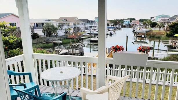 A covered outdoor area with a table and chairs, a view of a marina with boats, and a row of buildings in the background. - Cozy 3BR oceanfront home in Holden Beach, perfect for family getaways. (Holden Beach)
