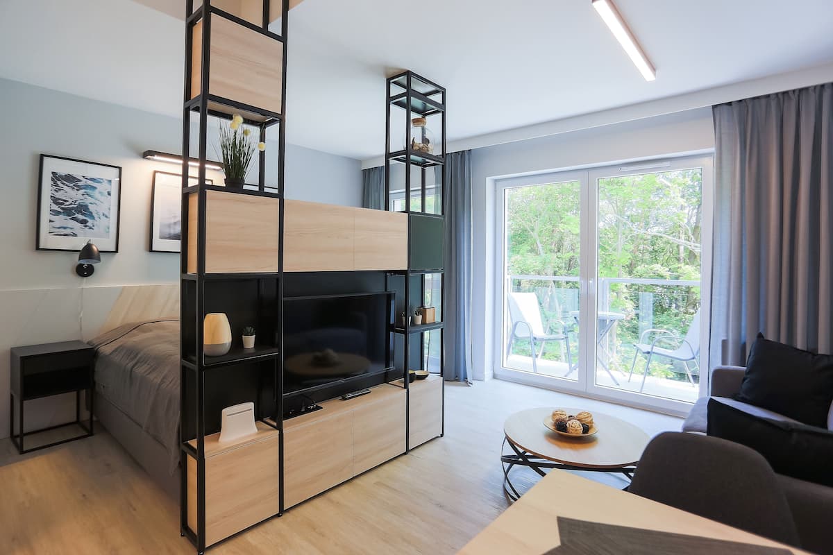Ein modernes Wohnzimmer mit Fernseher, Couchtisch und Sofa. Ein Balkon bietet Blick auf Grünflächen.