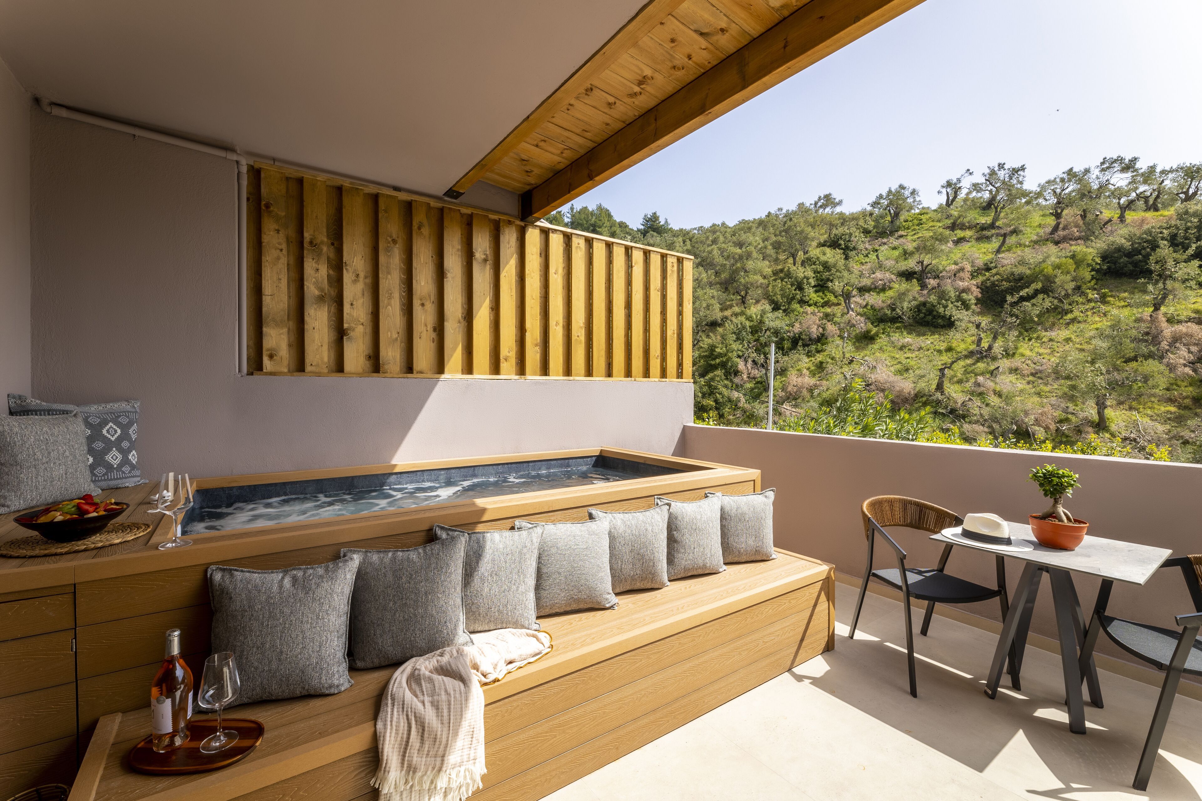 A wooden bench with cushions, a hot tub, and a table with chairs on a balcony overlooking a forested hillside.