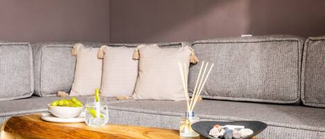 Ein modernes Wohnzimmer mit einem grauen Sofa, einem Holztisch, einer Schale grüner Trauben, einem Glas Wasser mit Zitrone und einer kleinen schwarzen Schale mit Snacks.