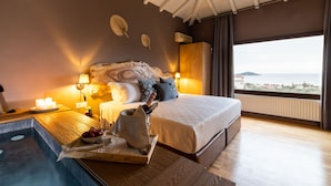 A bedroom with a large bed, a wooden nightstand, a small table with a bottle, a bowl of fruit, and a glass of wine. There is a window with a view of the sea and buildings. - Avaton Suites & Villa Philian Collection (Skiathos)