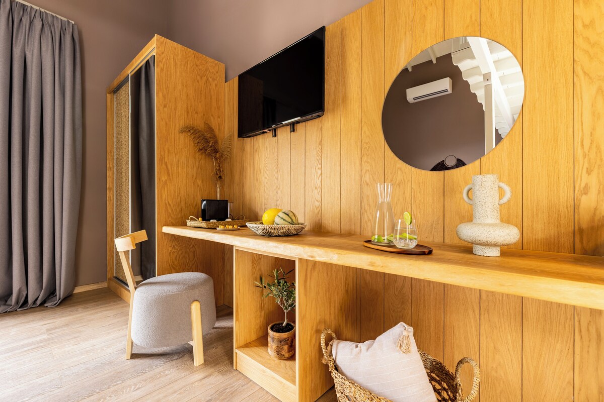A modern room with wooden panel walls, a flat-screen TV mounted above a wooden shelf, a small table with fruits and a vase, a chair, and a potted plant.