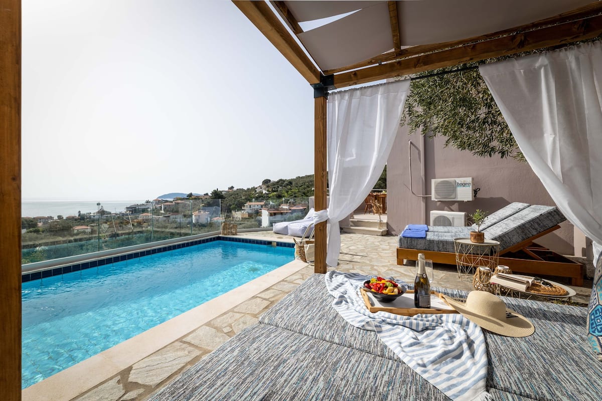 Un área de piscina en la azotea con vista al mar, una silla de descanso, una mesa con botella y fruta, y una pérgola con cortinas blancas.