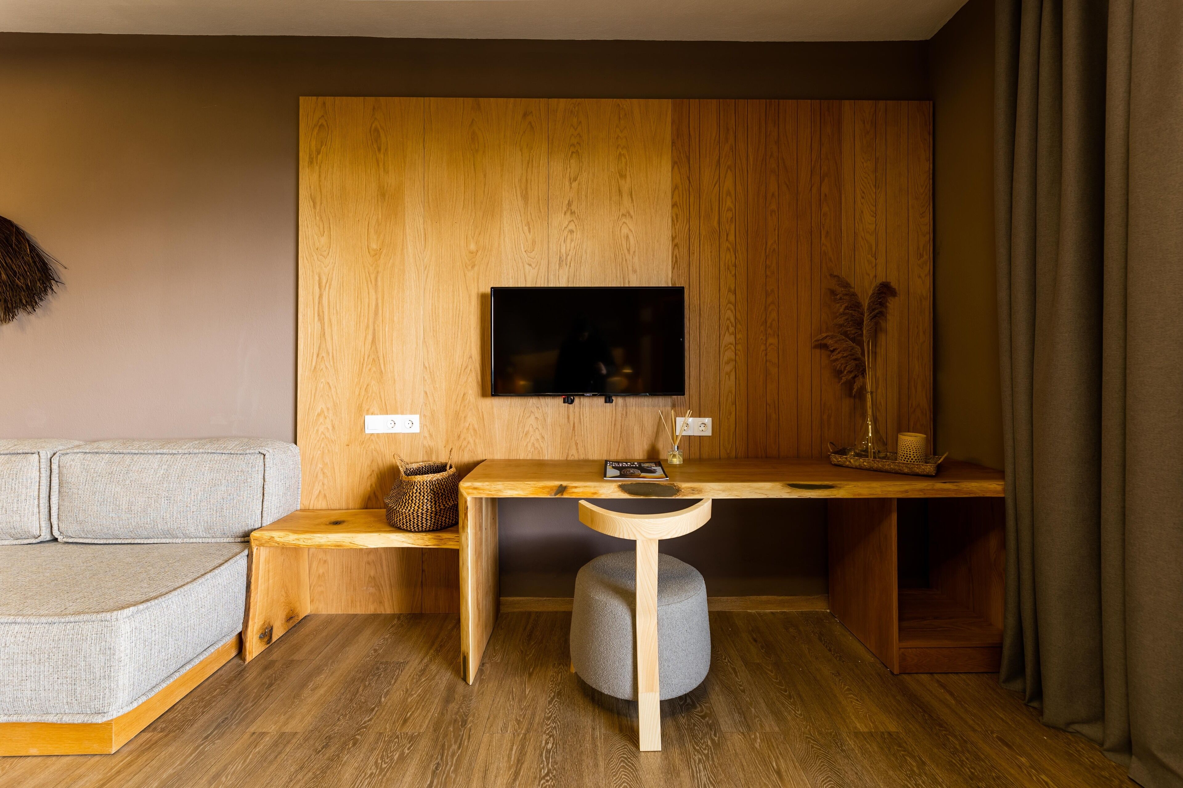 A modern living room with a wooden wall panel, a flat-screen TV mounted above a wooden desk, a grey sofa, and a small round ottoman.