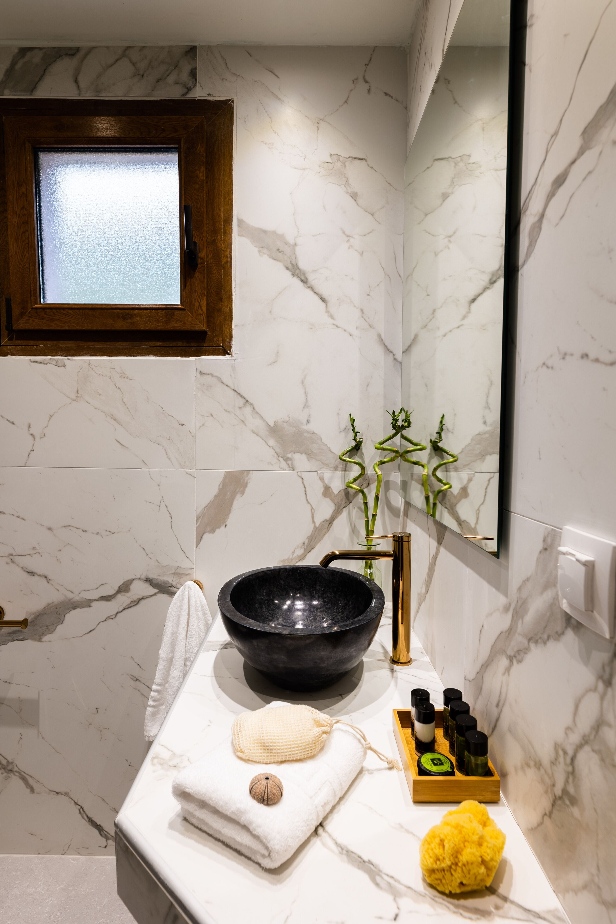 A bathroom with a marble wall, a wooden-framed window, a black basin, a towel, a yellow sponge, and a tray of toiletries.