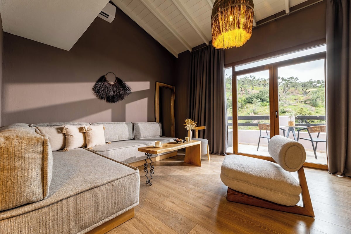 Una sala moderna con un sofá modular, una mesa de centro de madera y un sillón mecedor. Se ve un ventanal con vistas a la vegetación.