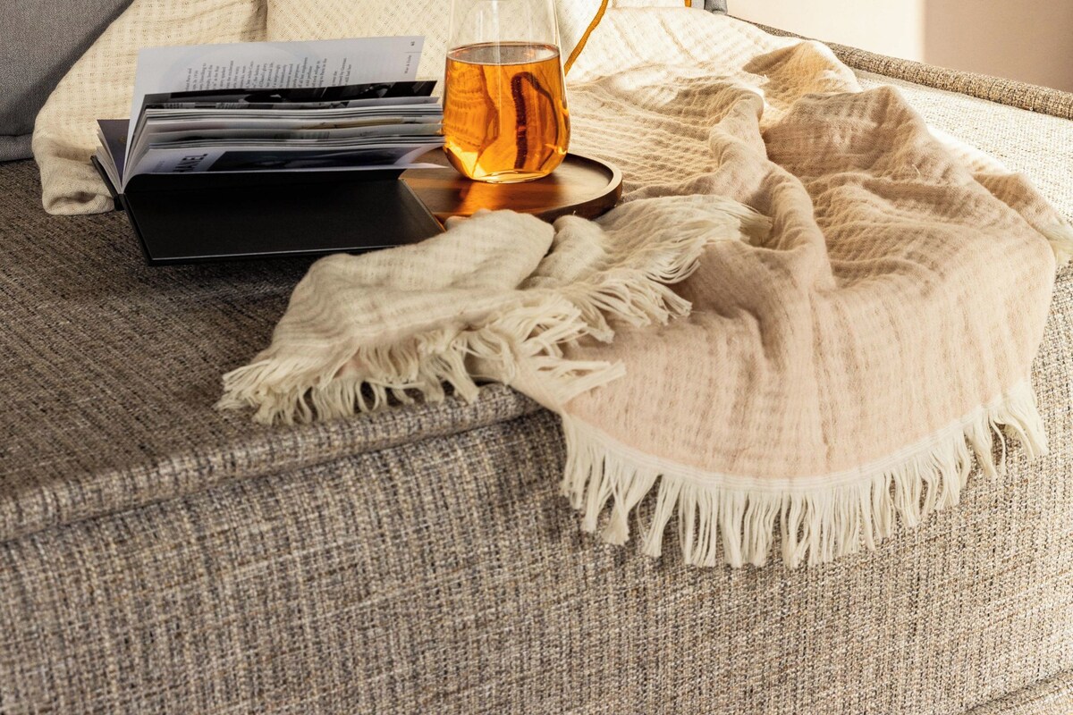 A bed with a blanket, a glass of amber liquid, and a book on a bedside table.