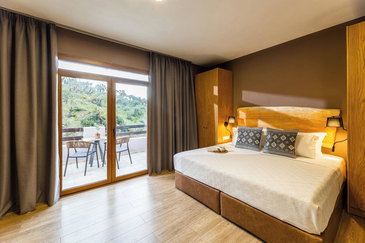Un dormitorio con una cama grande, cabecera de madera, balcón con sillas y vistas a zonas verdes.