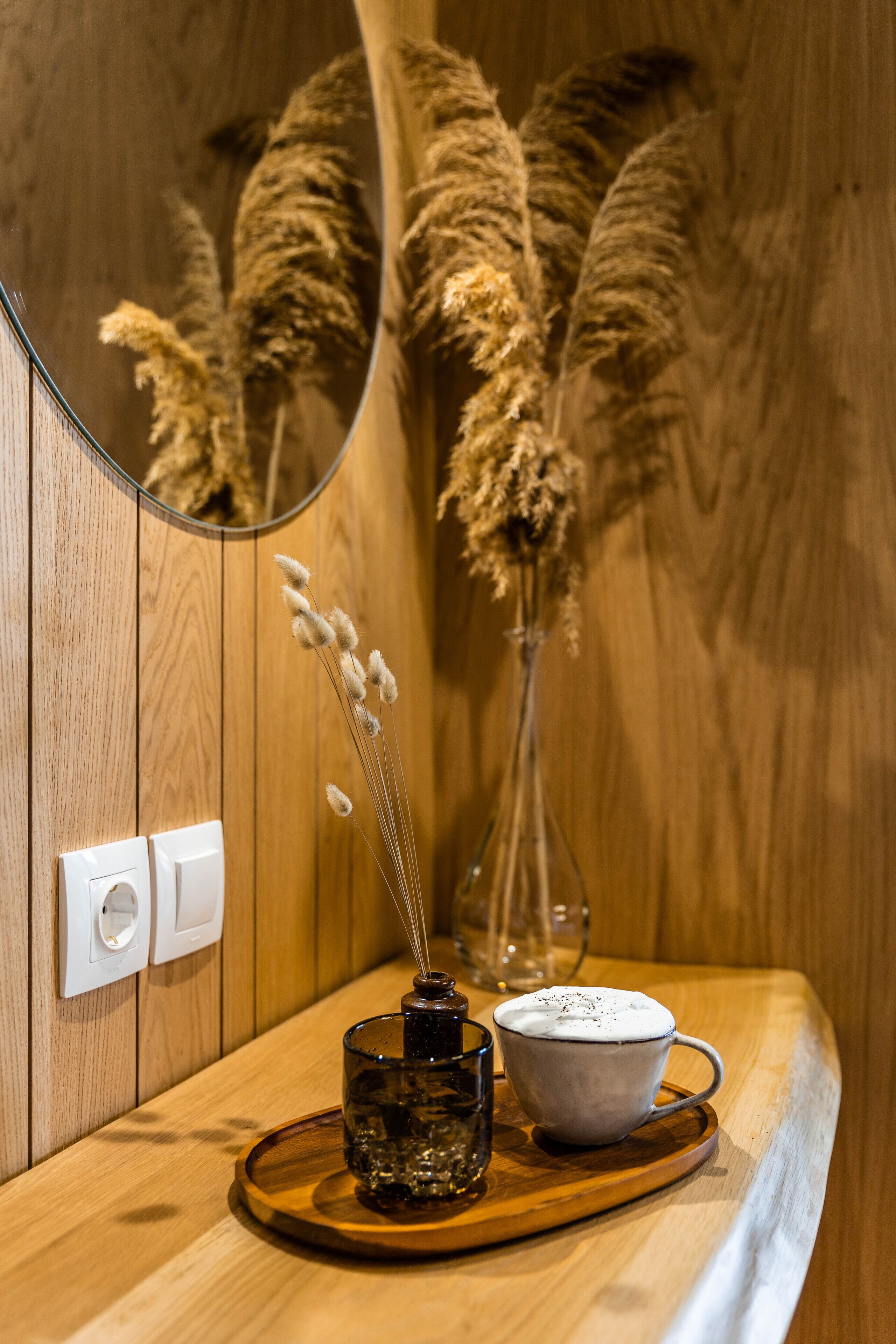 Ein Holzinterieur mit Spiegel, der Pampasgras reflektiert, einer Glasvase mit Pampasgras, einem Glas Wasser und einer weißen Tasse auf einem Holztablett.