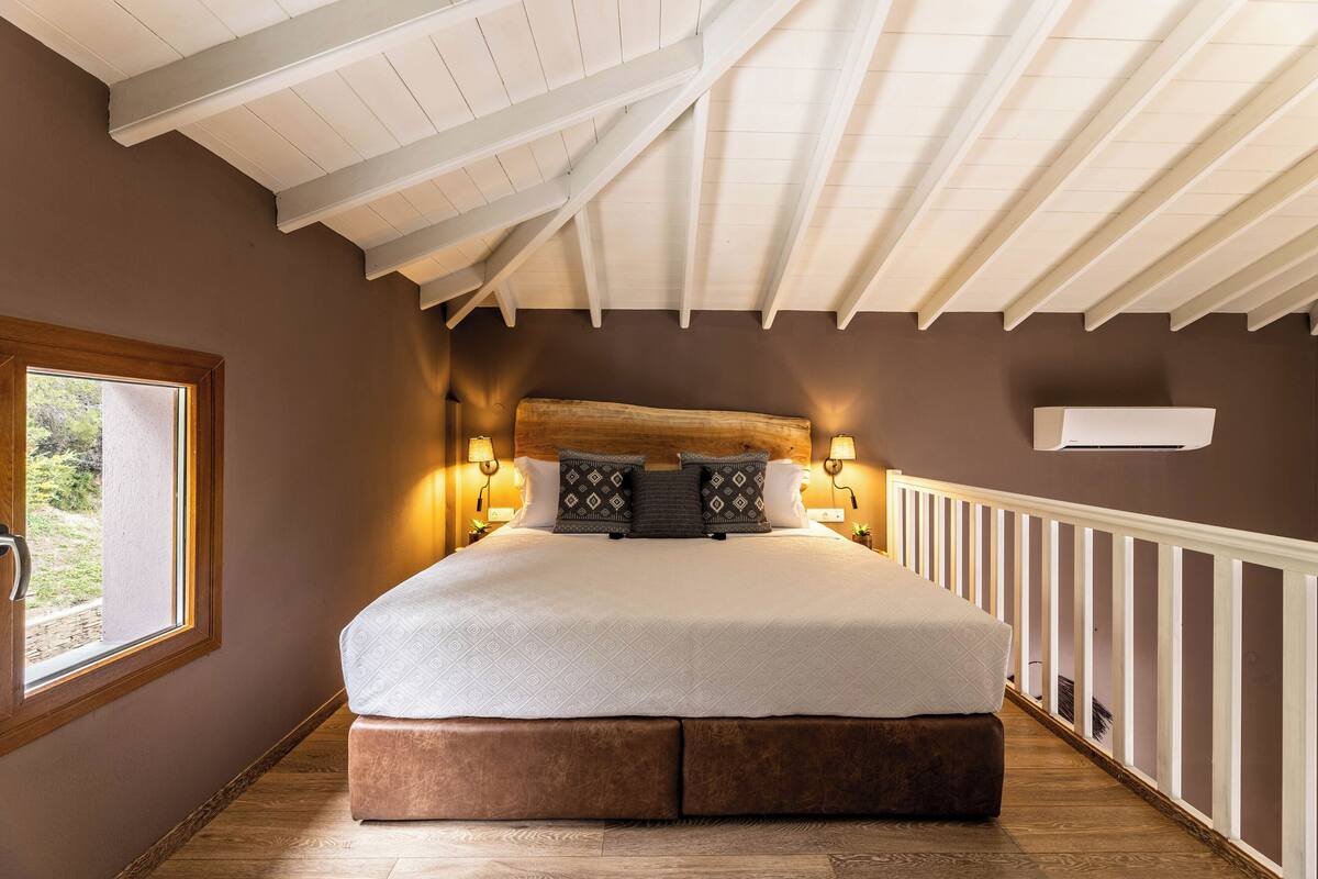 Un dormitorio con una cama grande, cabecera de madera, dos lámparas fijadas a la pared, una ventana y una barandilla blanca.