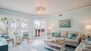 A spacious living room with a large window, a white sofa with teal pillows, a coffee table, and a dining area with chairs and a table.