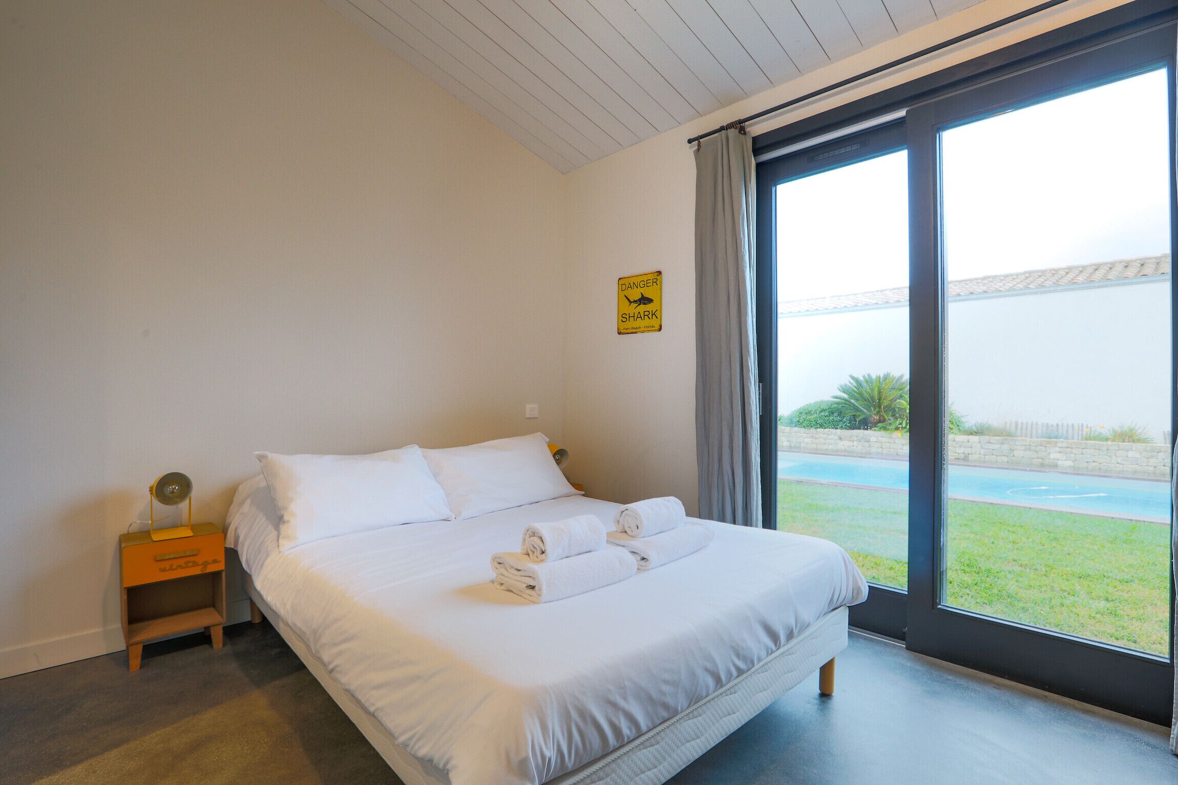 Une chambre avec un lit, une table de chevet, une fenêtre donnant sur une piscine et de la verdure, et un panneau « Attention, requin » accroché au mur.