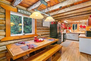 A log cabin interior with a wooden dining table, bench, and a window with a view of the forest. - Escape to 5 Private Acres | River Access, Hot Tub & Wildlife Watching (Leavenworth)