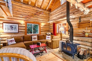 A log cabin interior with a sofa set, a red coffee table, a wood-burning stove, and framed pictures on the walls. - Escape to 5 Private Acres | River Access, Hot Tub & Wildlife Watching (Leavenworth)