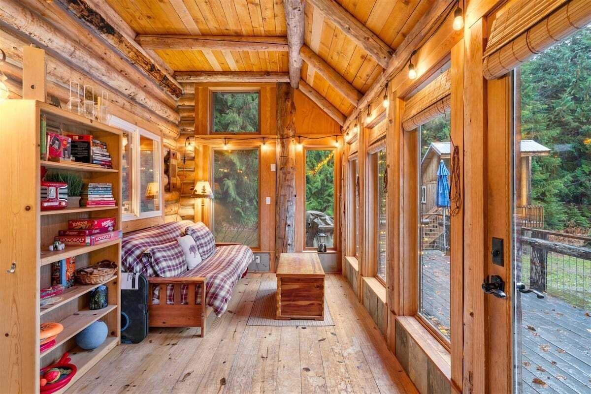 A cozy log cabin interior with a bed, shelves with books, and large windows offering a view of the outdoors.