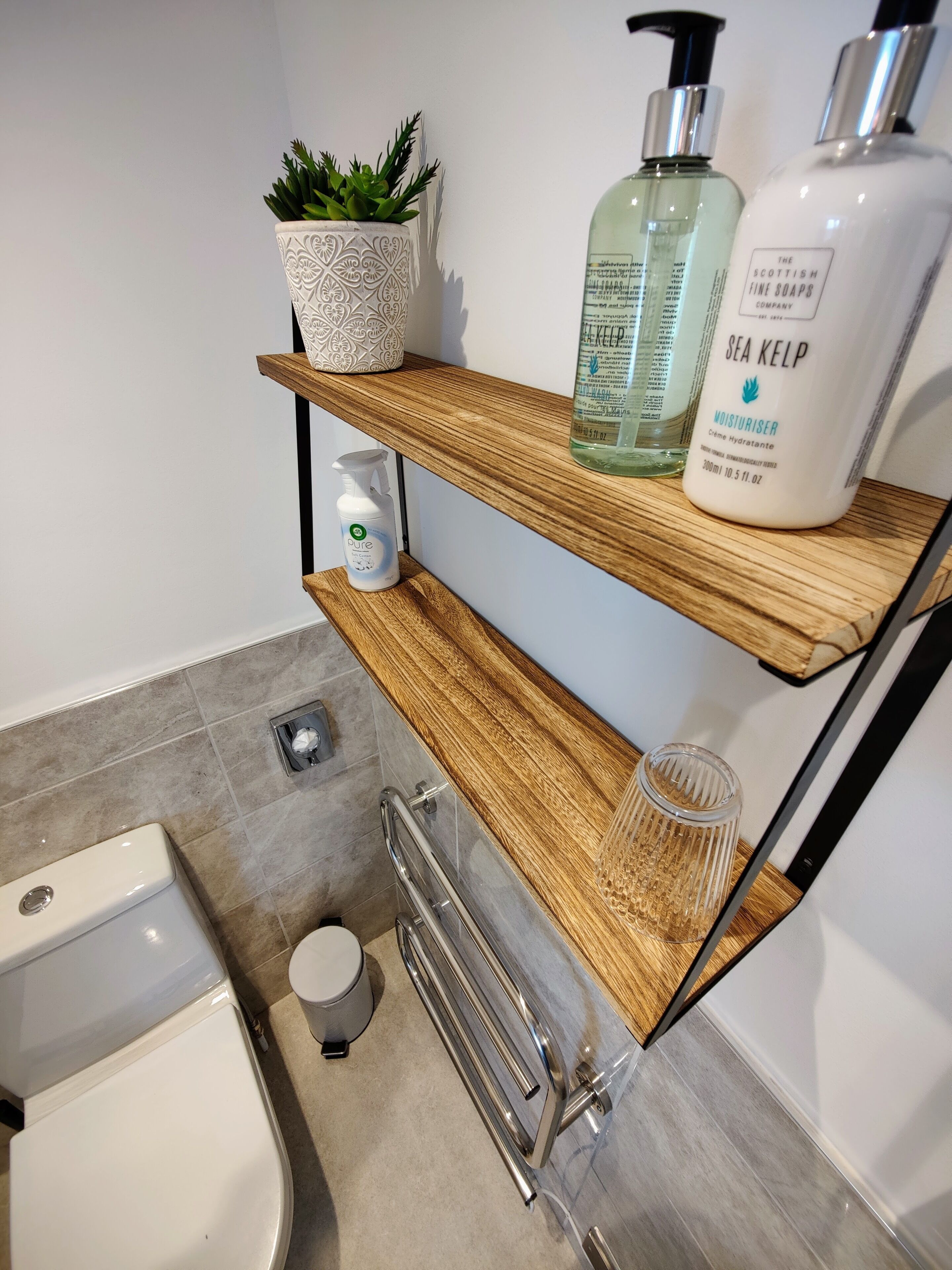 Un baño con un estante de madera que sostiene una planta en maceta, un frasco de crema hidratante de alga marina y un recipiente de vidrio.
