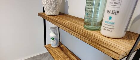 Un baño con un estante de madera que sostiene una planta en maceta, un frasco de crema hidratante de alga marina y un recipiente de vidrio.