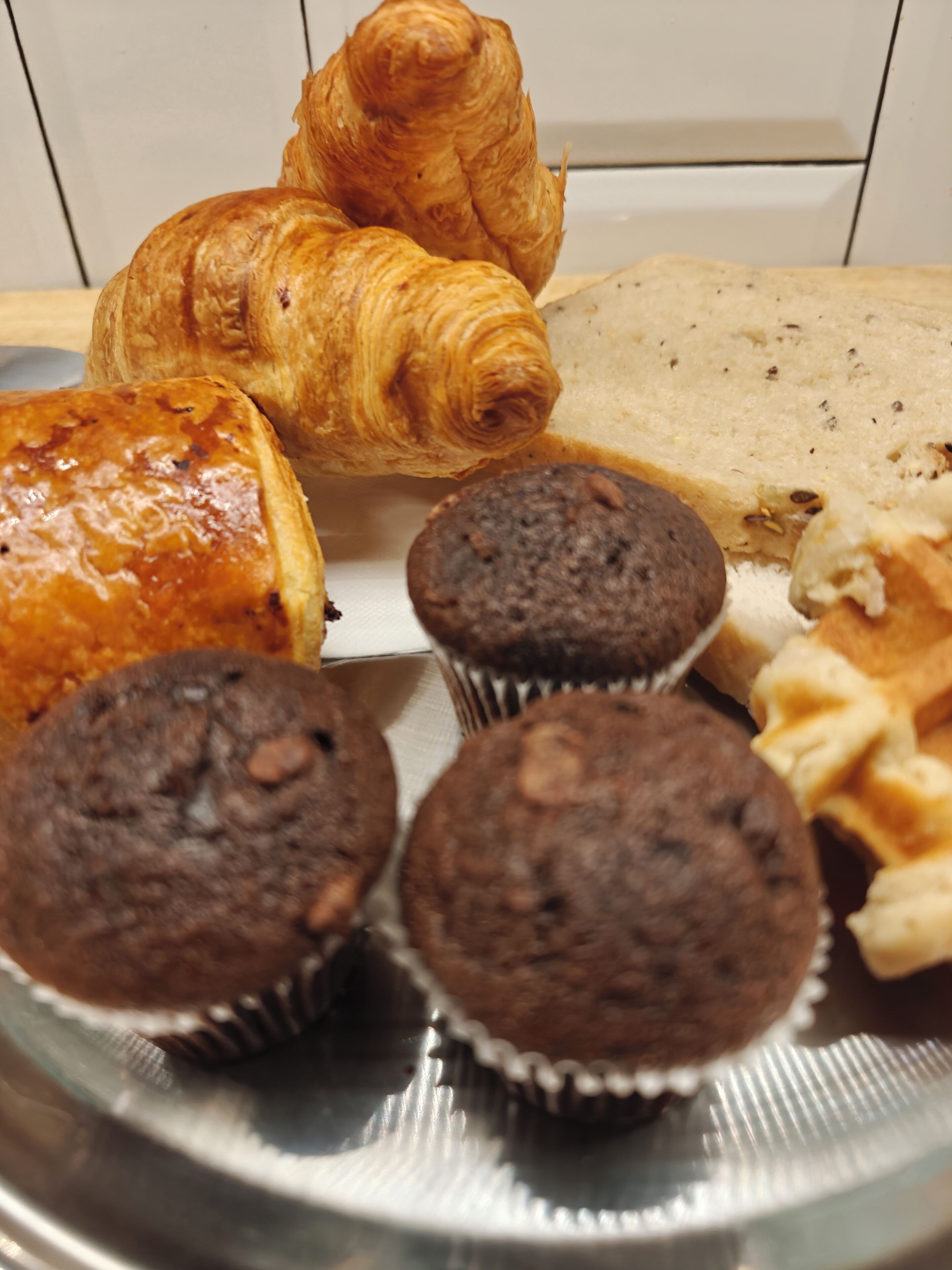 Diversos produtos de panificação em uma bandeja, incluindo croissants, muffins e uma barra de pão.