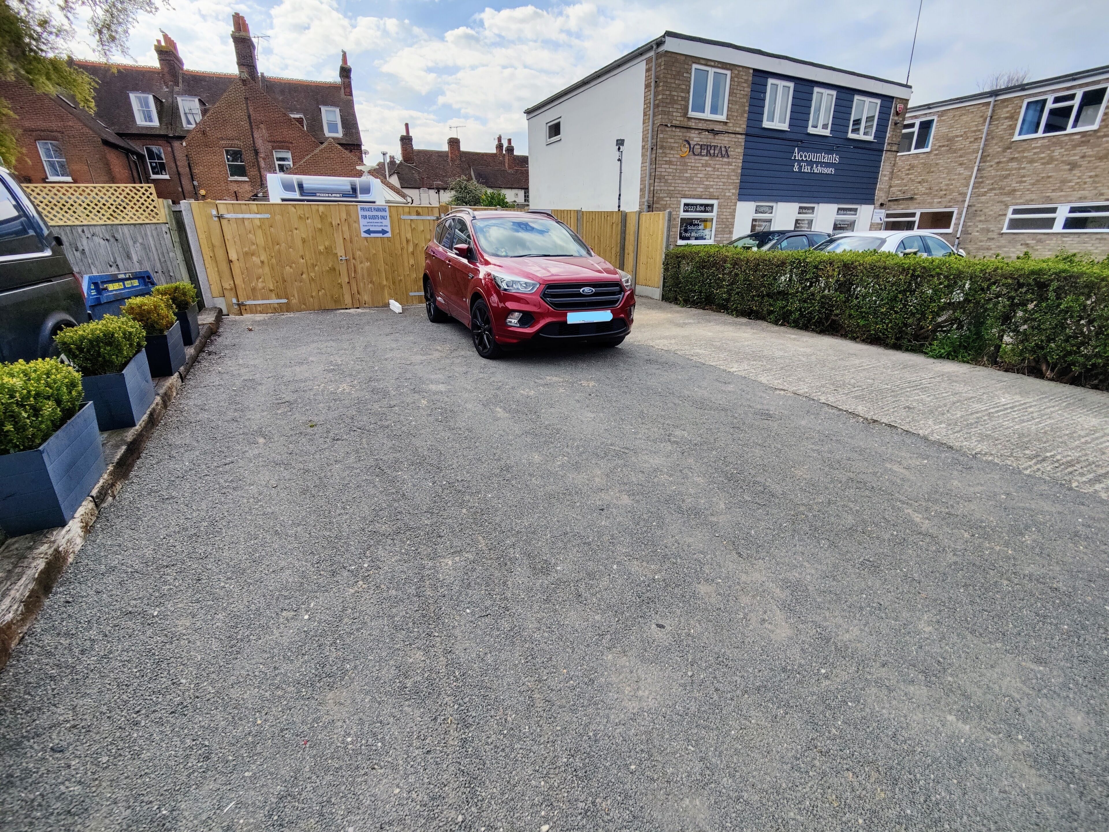 A red car parked in a driveway with a wooden gate and a sign indicating private parking for guests only.