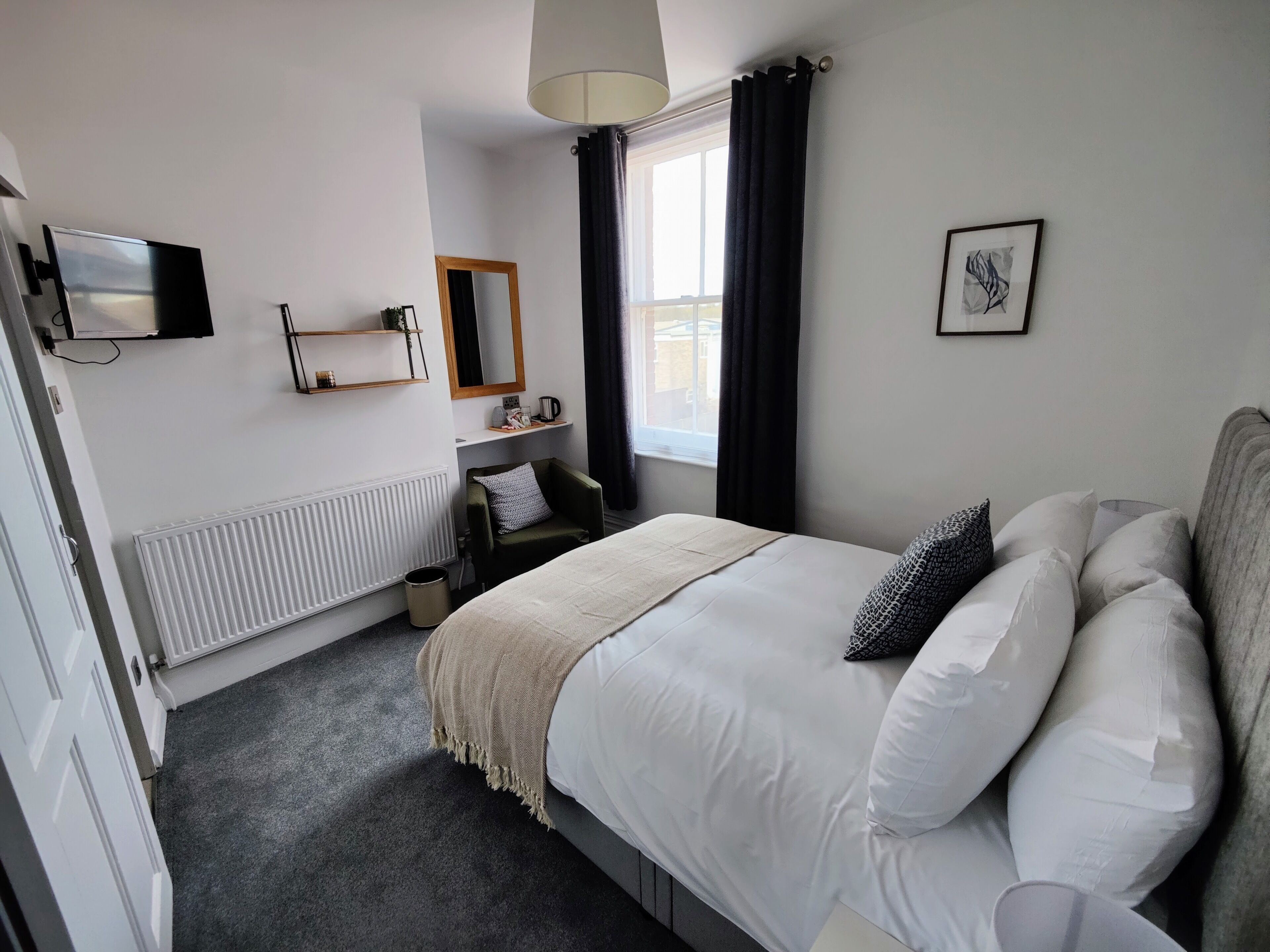 Een moderne hotelkamer met een groot bed, een aan de muur bevestigde televisie, een raam met gordijnen, een radiator en een klein plankje met spullen.