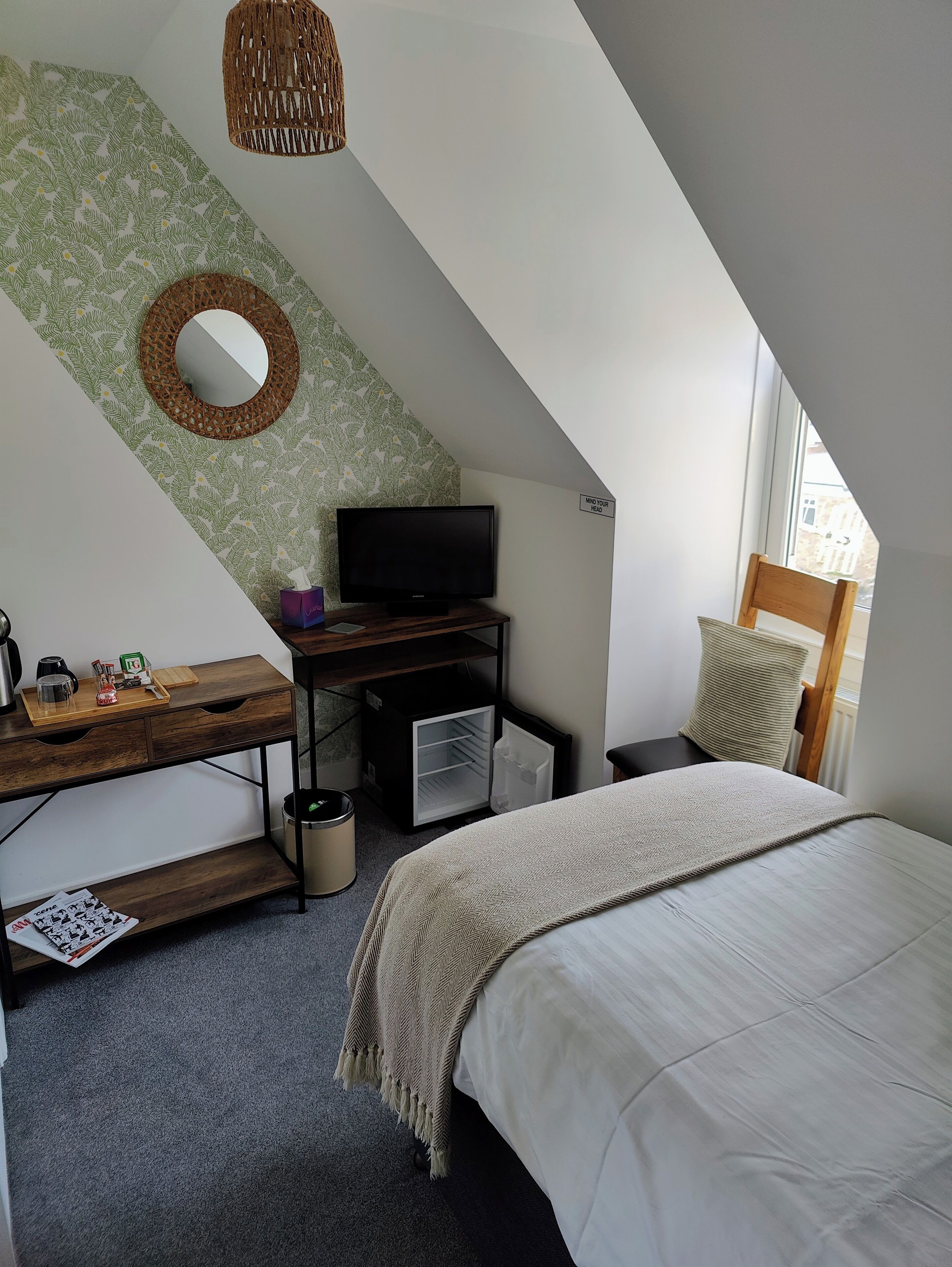 A small, modern bedroom with a sloped ceiling, a bed, a desk with a television, a small refrigerator, and a window with a view of buildings outside.