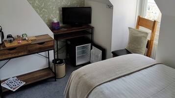 A small, modern bedroom with a sloped ceiling, a bed, a desk with a television, a small refrigerator, and a window with a view of buildings outside.