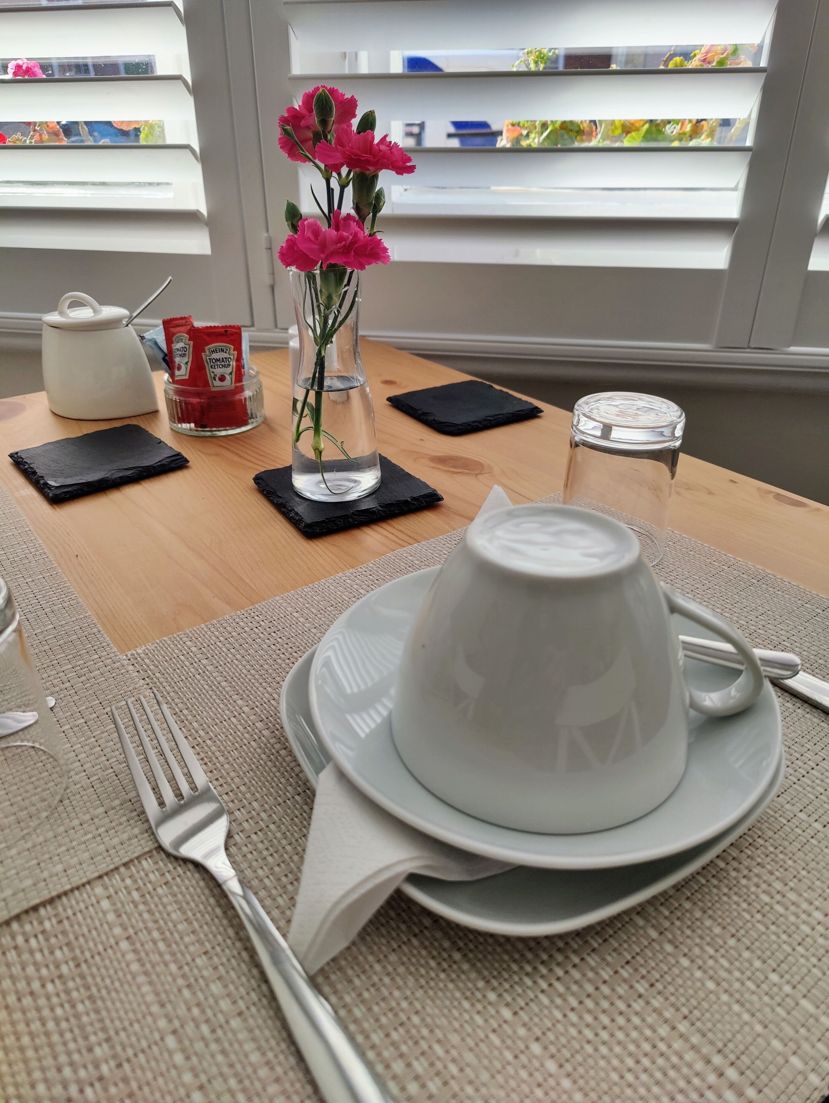 Mesa posta com um bule branco, uma chávena branca, um garfo e um guardanapo. Há um vaso com flores cor-de-rosa e um copo de água sobre a mesa.